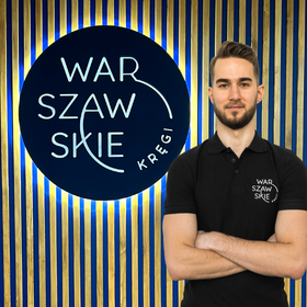 Fizjoterapeuta gabinetu Warszawskie Kręgi Maciej Szumielewicz ubrany w granatową koszulkę firmową na tle firmowego logo.