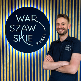 Fizjoterapeuta gabinetu Warszawskie Kręgi Maciej Berond ubrany w granatową koszulkę firmową na tle firmowego logo.