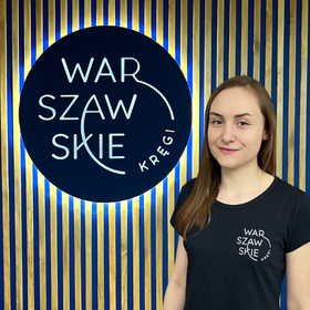 Fizjoterapeutka gabinetu Warszawskie Kręgi Iga Wróblewska ubrana w granatową koszulkę firmową na tle firmowego logo.