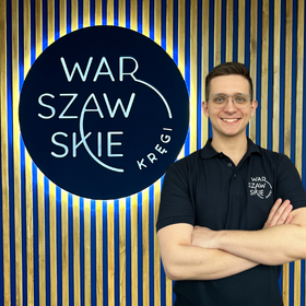 Fizjoterapeuta gabinetu Warszawskie Kręgi Jakub Kozłow ubrany w granatową koszulkę firmową na tle firmowego logo.