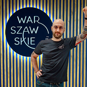 Fizjoterapeuta i założyciel gabinetu Warszawskie Kręgi Patryk Wąsowski ubrany w granatową koszulkę firmową na tle firmowego logo.
