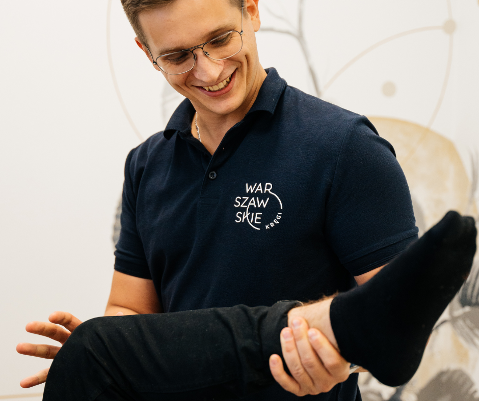 Młody i uśmiechnięty fizjoterapeuta w okularach i koszulce z logo gabinetu fizjoterapii Warszawskie Kręgi przytrzymuje nogę pacjenta testując zakresy ruchu.